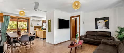 Una sala moderna con un sofá de cuero marrón, un comedor con mesa y sillas de madera, y una cocina al fondo.