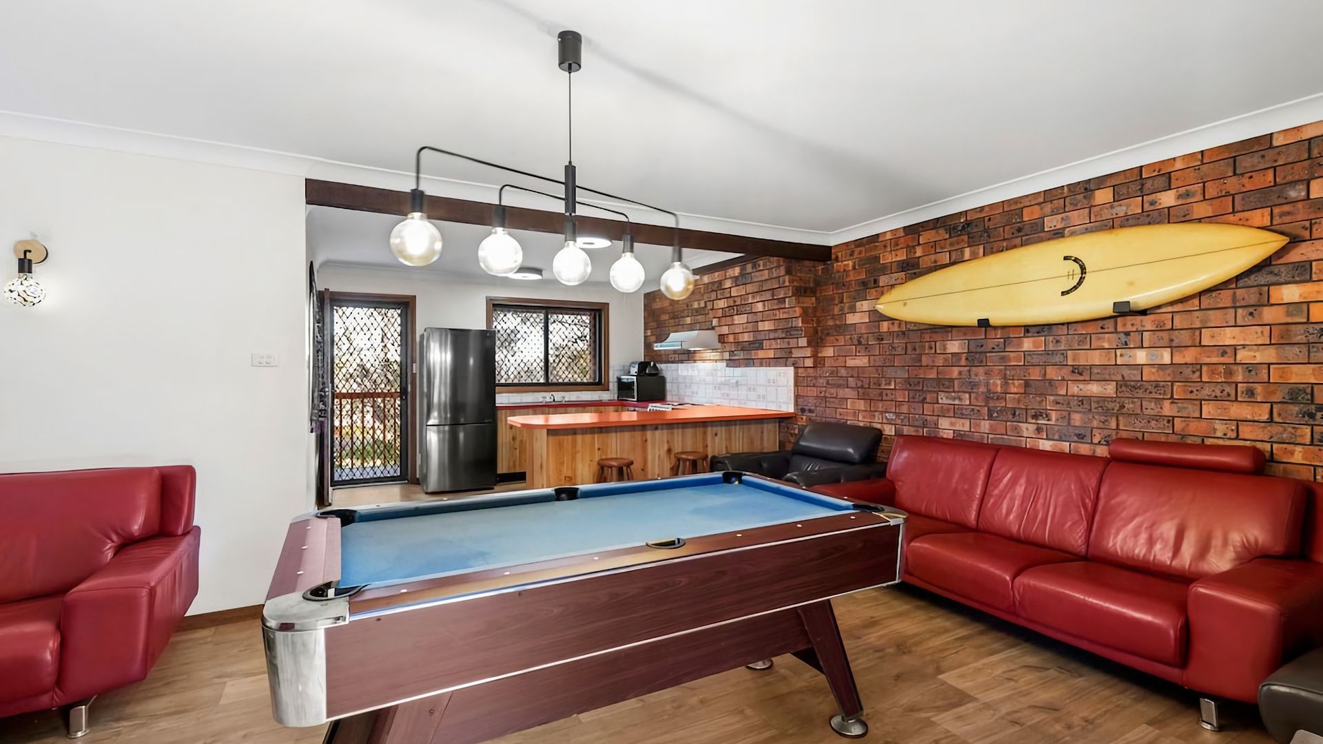 Een pooltafel in een woonruimte met rode leren banken, een bakstenen muur en een surfplank.