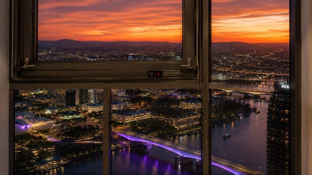 Una vista panorámica de un paisaje urbano al atardecer, vista a través de una ventana que muestra el horizonte de la ciudad, un río y un puente.