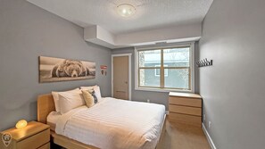 Un dormitorio con cama, mesita de noche, cómoda, ventana y un cuadro de un oso en la pared.