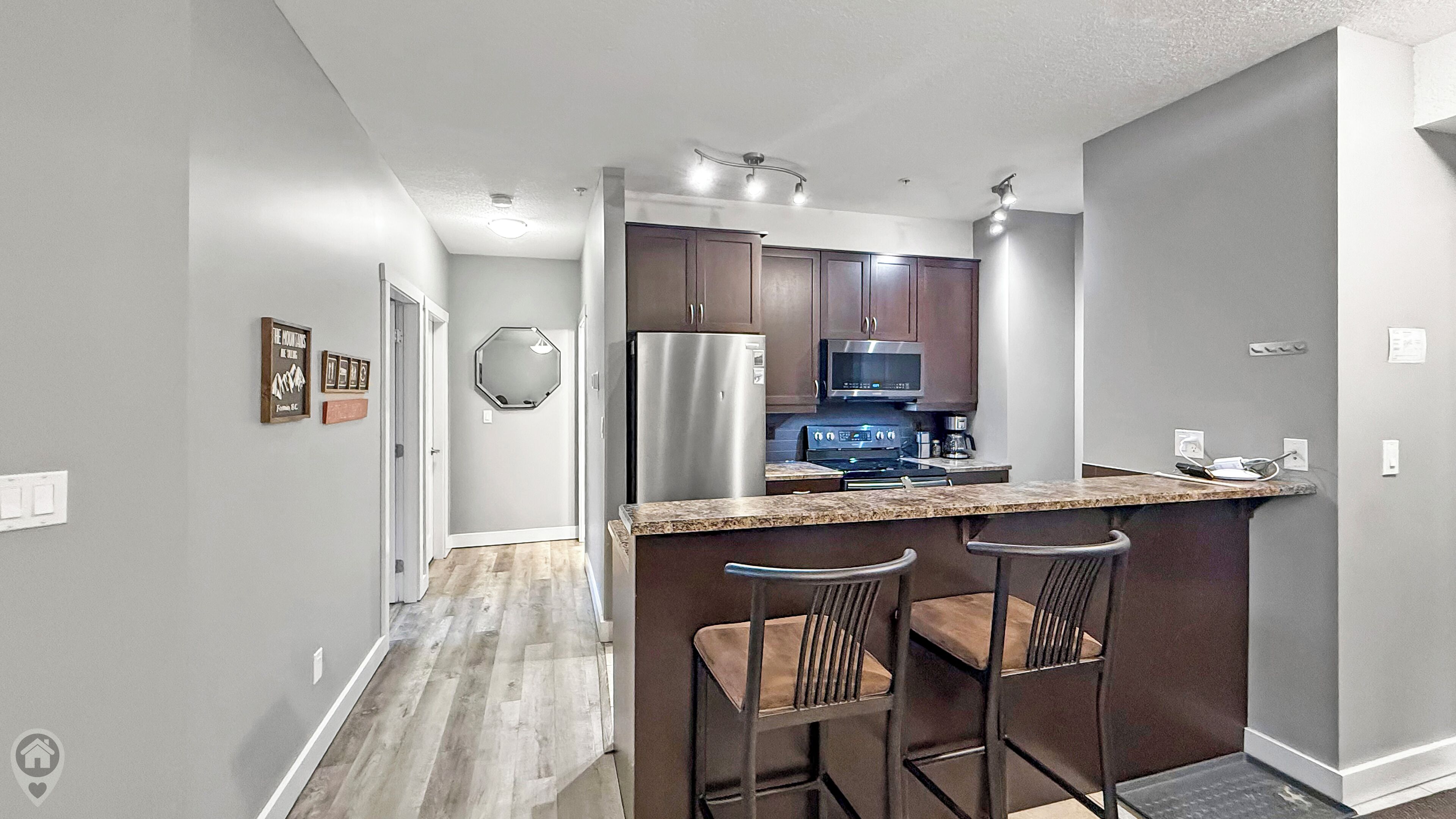 A modern kitchen with dark wooden cabinets, stainless steel appliances, and a granite countertop. There are two bar stools with cushioned seats.