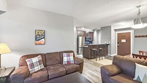 A modern living room with a sofa and armchair, a kitchen in the background, and a sign on the wall.