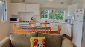 A compact kitchen with white cabinets, a dining table with chairs, a microwave, a stove, and a refrigerator. - Sting Ray Bay near Boat Harbour Beach (Bendalong)