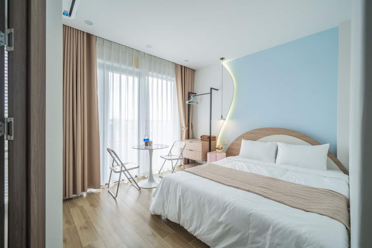 A hotel room with a bed, bedside table, lamp, and a view of the outdoors through large windows.