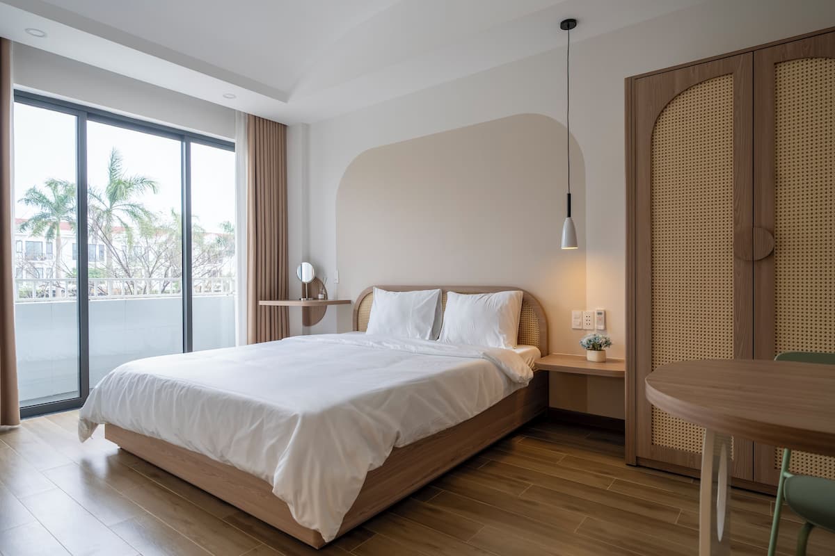 A modern hotel room with a large bed, a bedside table with a lamp, a small table, and a view of palm trees through large windows.