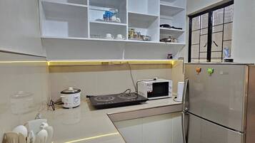 A compact kitchen with white cabinets, a stainless steel refrigerator, a microwave, and a stove. There are shelves above the counter and a window with blinds.