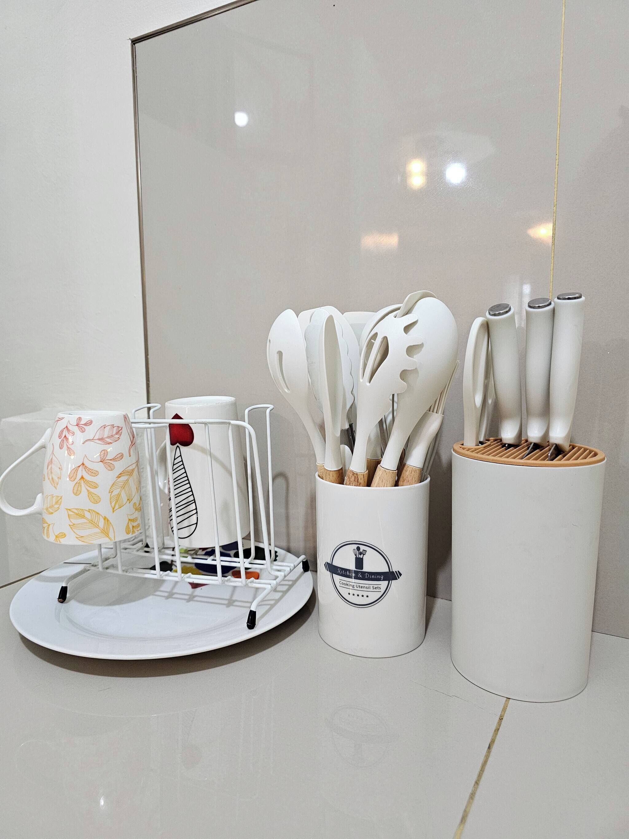 A kitchen area with a white mug with leaf patterns, a white utensil holder with a spoon, and a white container with wooden utensils.