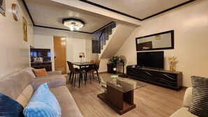 A modern living room with a sofa, coffee table, and TV. There's a dining area with chairs and a table. A staircase leads to an upper level. - Cozy Biñan Getaway - Entire House for Rent (Biñan City)
