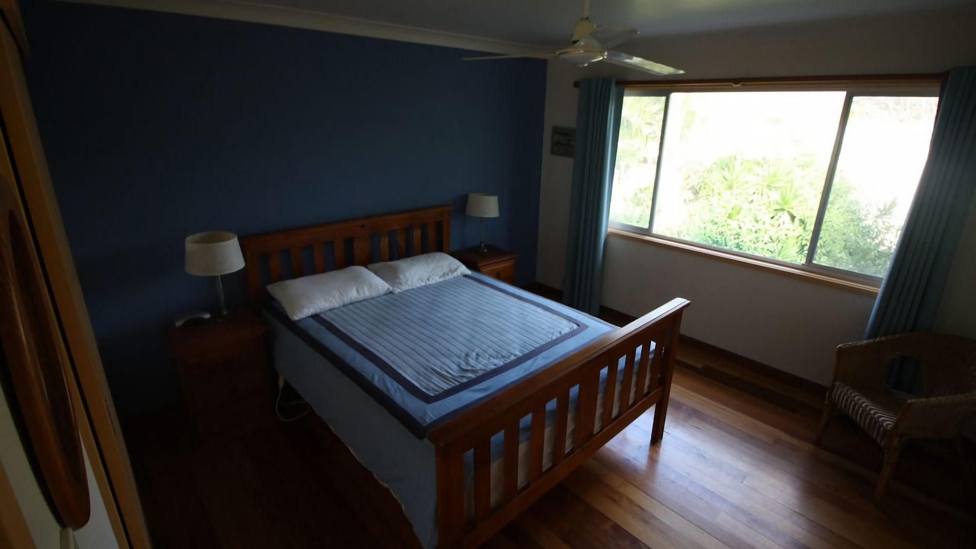 A bedroom with a wooden bed, a nightstand with a lamp, a window with curtains, and a chair.