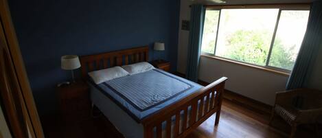 A bedroom with a wooden bed, a nightstand with a lamp, a window with curtains, and a chair.