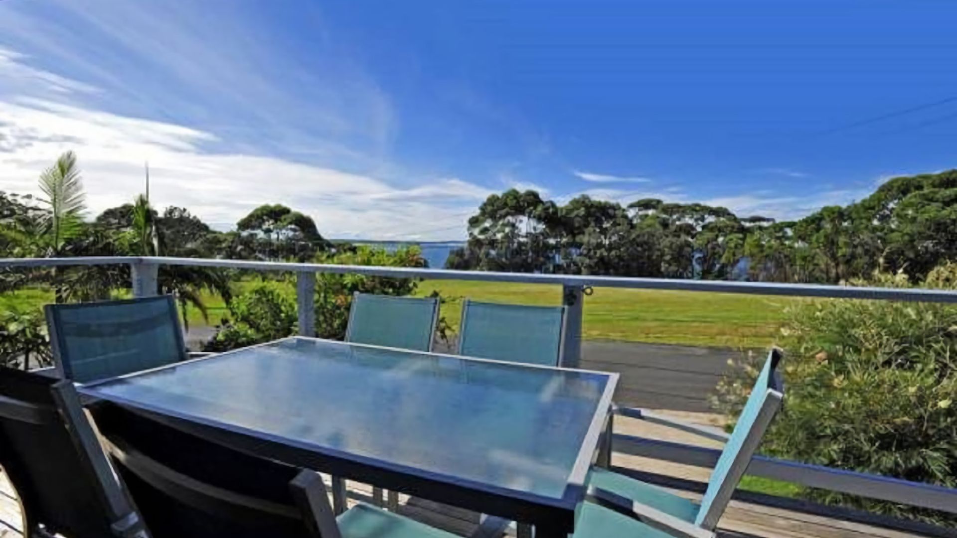 A balcony with a glass table and chairs, overlooking a grassy area and trees.