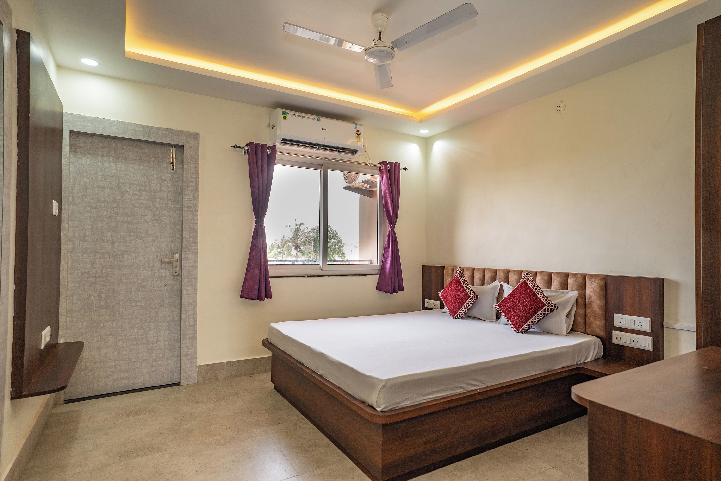 A bedroom with a bed, a ceiling fan, an air conditioning unit, a window with curtains, and a door.