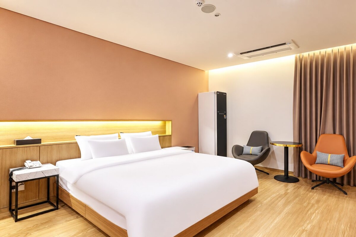 A hotel room with a large bed, bedside table with a phone, a chair, a desk, and a large window with curtains.