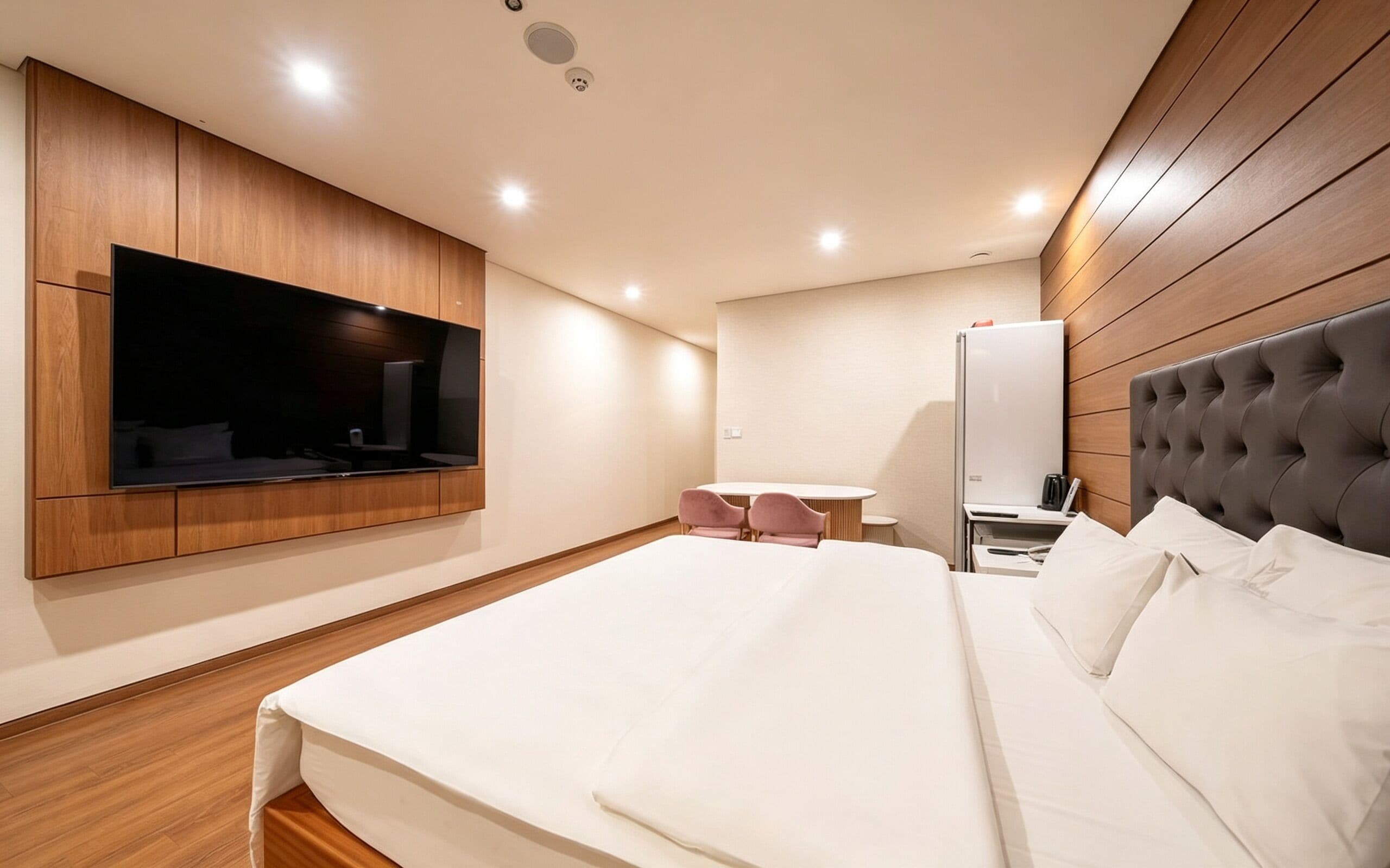 A modern hotel room with a large bed, a flat-screen TV mounted on the wall, a small round table with chairs, and a bedside table with a lamp.
