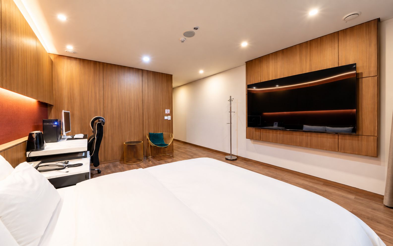 A modern hotel room with a large bed, a desk with a computer, a chair, a small table, and a TV mounted on the wall.