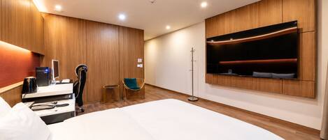 A modern hotel room with a large bed, a desk with a computer, a chair, a small table, and a TV mounted on the wall.