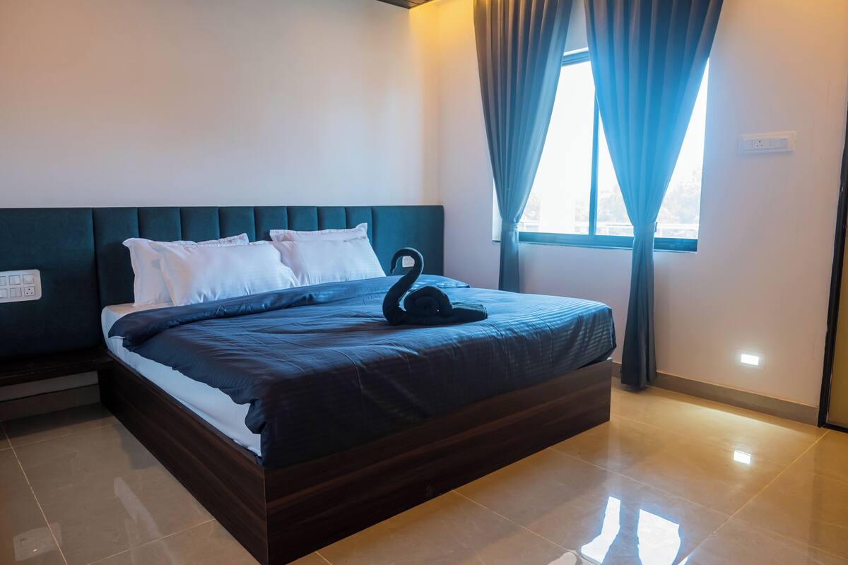 A neatly made bed with a dark headboard and white pillows, a swan-shaped pillow, and a window with curtains.