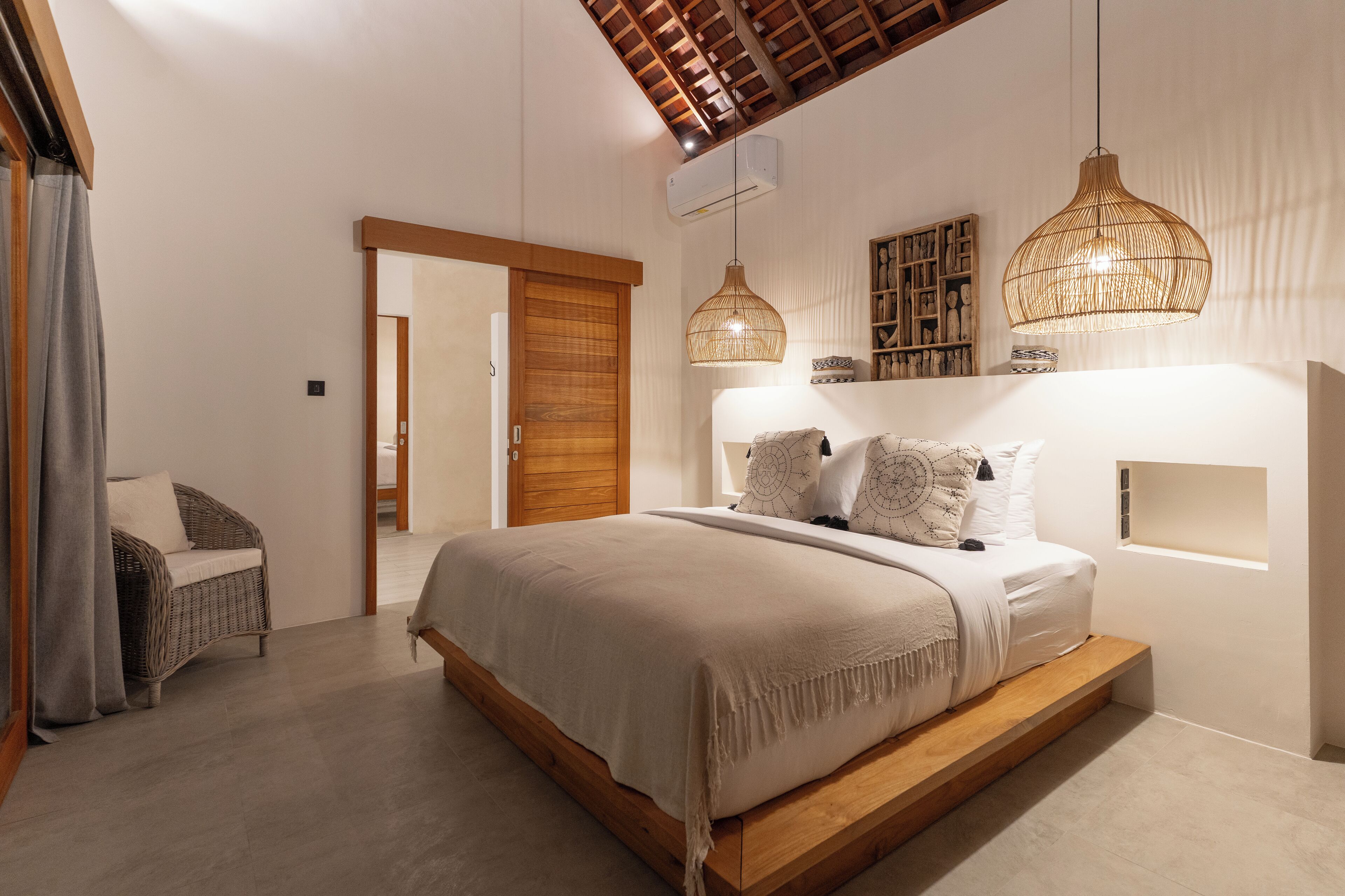 A bedroom with a large bed, wooden headboard, two hanging light fixtures, a built-in shelf, and a sliding door leading to another room.
