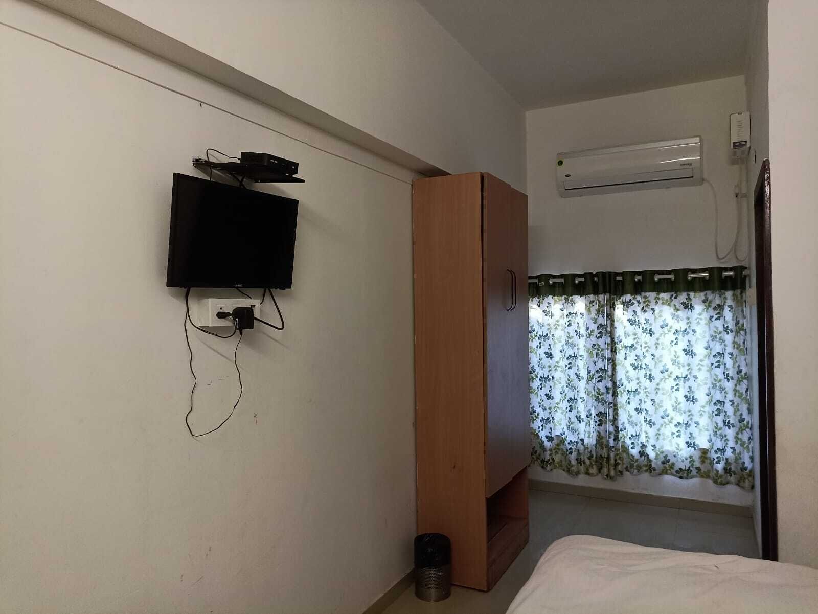 A bedroom with a bed, a TV mounted on the wall, a wardrobe, and a window with floral curtains.