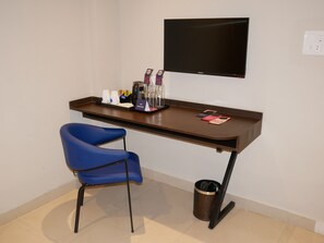 A small, clean room with a desk, a blue chair, a TV, and a trash can. - Surendra Landmark By Spree Hotels (Bhopal)