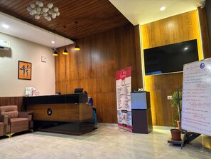 A hotel reception area with a wooden reception desk, a seating area with a sofa, a wall-mounted air conditioner, and a digital screen. - Surendra Landmark By Spree Hotels (Bhopal)