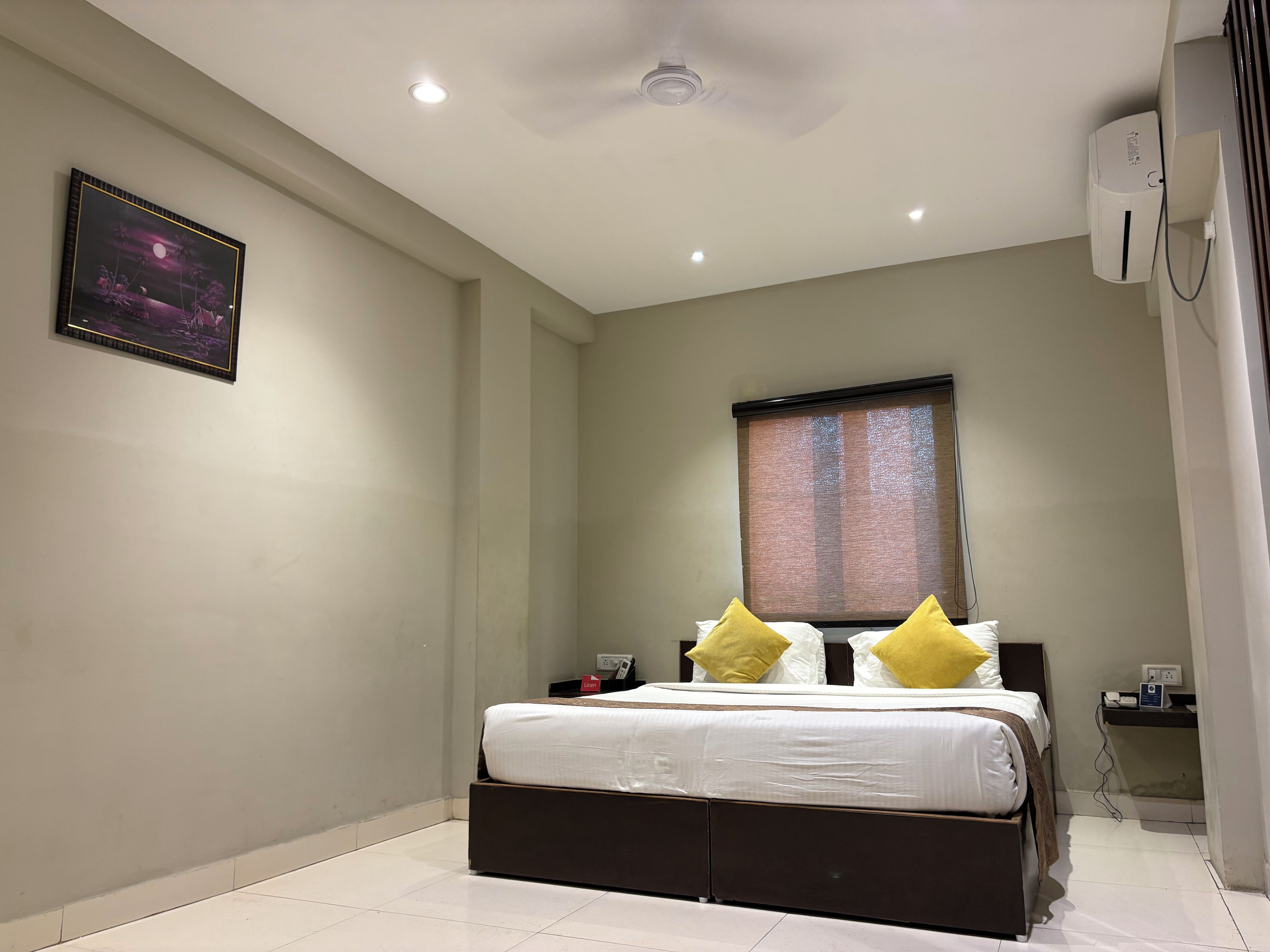 A hotel room with a bed, a nightstand, a wall-mounted air conditioner, and a window with blinds.