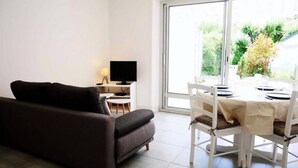 Una sala de estar moderna con un sofá, televisor, mesa de comedor con sillas y un amplio ventanal con vista a vegetación.