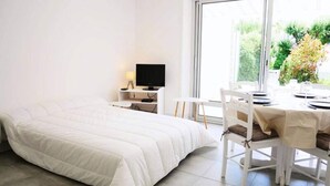 Habitación de hotel con una cama grande, un televisor sobre un mueble, una mesa de comedor con sillas y vistas a zonas verdes.