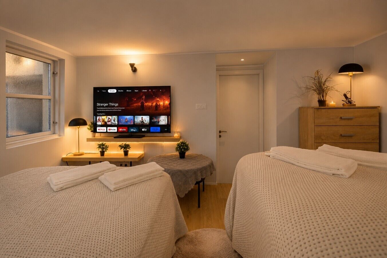 A hotel room with two beds, a TV displaying a movie, a bedside table with a lamp, and a wooden dresser.