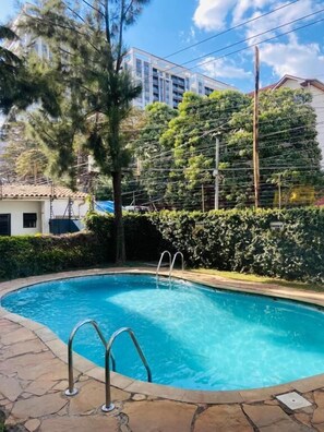 A swimming pool with a metal ladder, surrounded by stone tiles, with trees and a building in the background. - Riara Prestige Apartments (Nairobi)