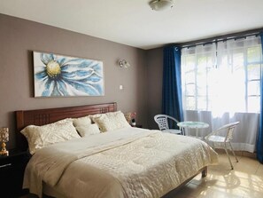 A bedroom with a bed, a bedside table, a lamp, a chair, a small table, and a window with curtains. - Riara Prestige Apartments (Nairobi)