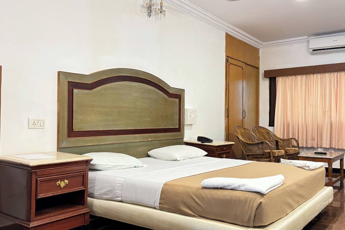 Una camera d'albergo con un letto grande, un comodino in legno, una scrivania con una sedia, un condizionatore d'aria e una finestra con tende.