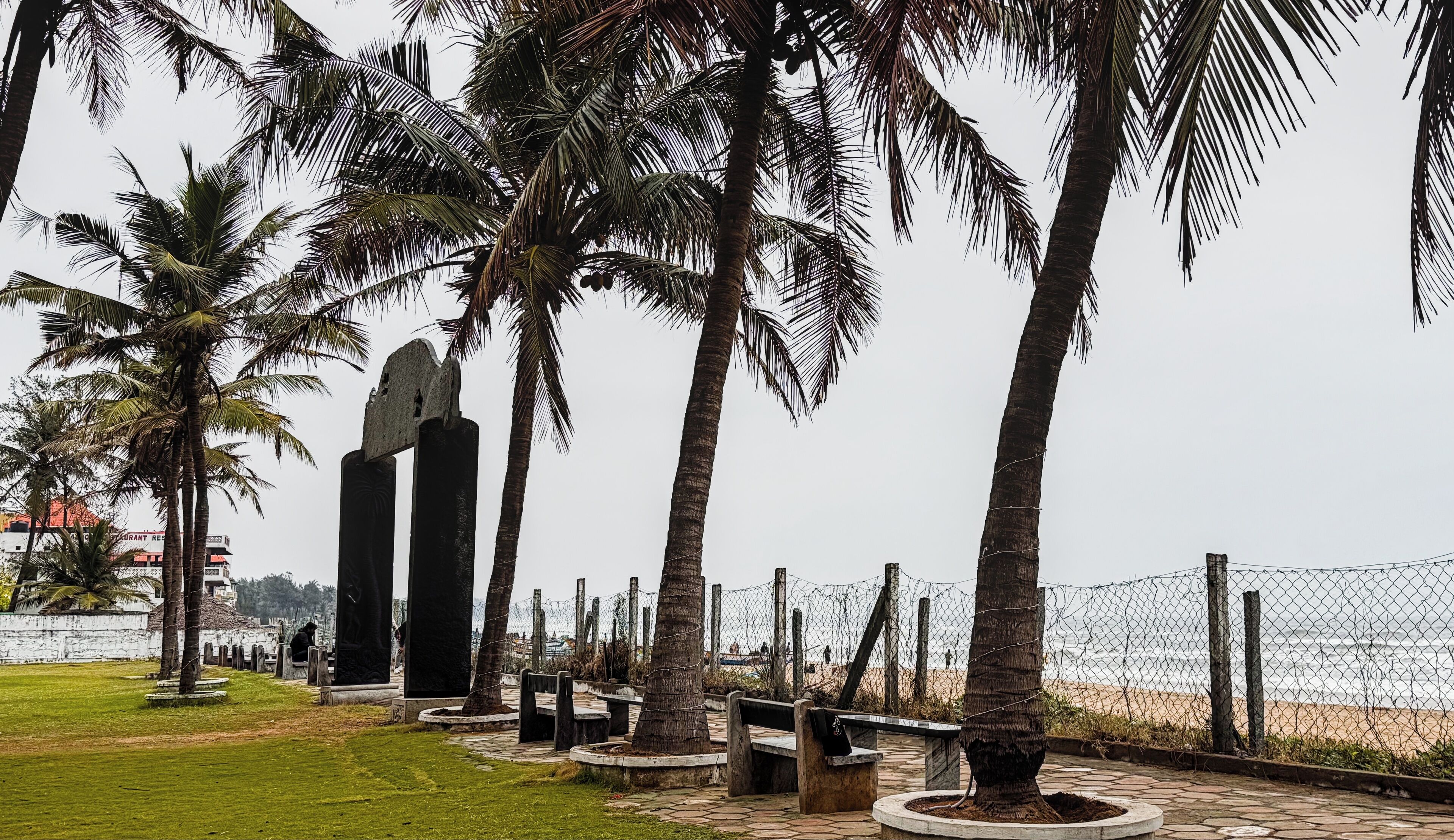 A coastal walkway with palm trees, stone benches, and a fence overlooking the beach.