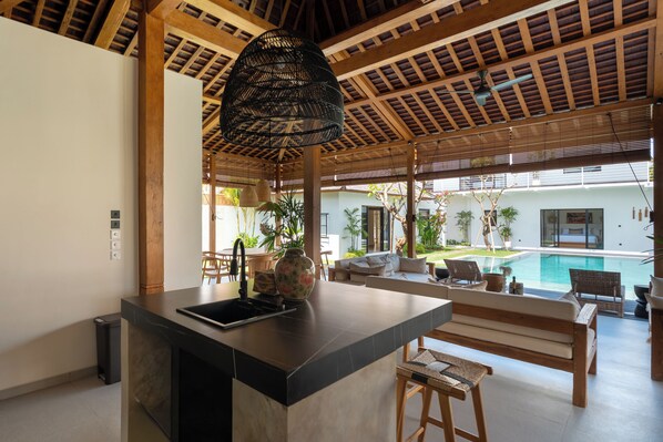 A modern kitchen with a large island, wooden ceiling beams, and a view of a pool and outdoor seating area. - Villa Nador by Alfred in Bali (Seminyak)