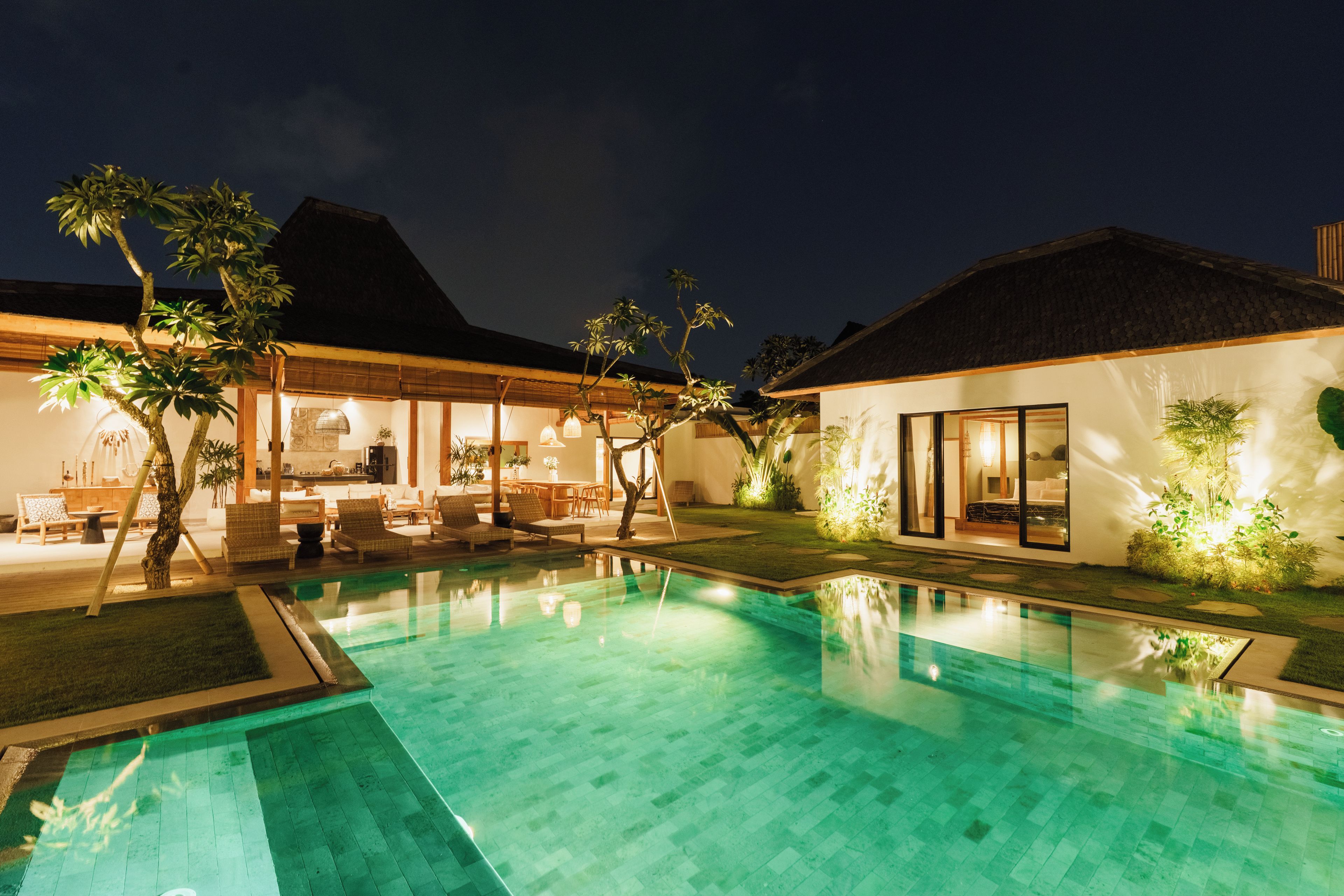 A nighttime view of a resort with a swimming pool, lounge area, and a thatched-roof building.