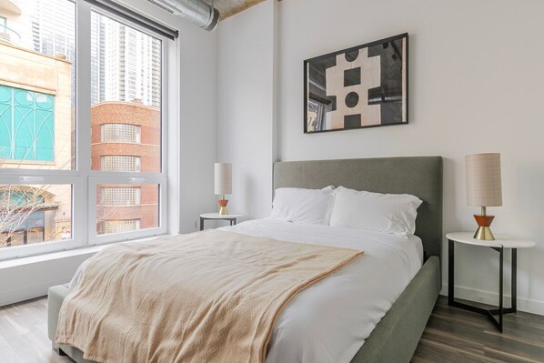 Une chambre d’hôtel moderne dotée d’un grand lit, de deux tables de chevet, d’une fenêtre donnant sur la ville et d’une œuvre d’art abstraite accrochée au mur.