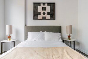 Un lit bien fait, avec une tête de lit grise et du linge de lit blanc, flanqué de deux tables de chevet surmontées de lampes, dans une chambre minimaliste avec un mur uni et une œuvre d'art abstraque encadrée au-dessus du lit.