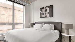 Ein modernes Hotelzimmer mit einem großen Bett, zwei Nachttischlampen, einem an der Wand befestigten abstrakten Kunstwerk und einem großen Fenster mit Blick hinaus.