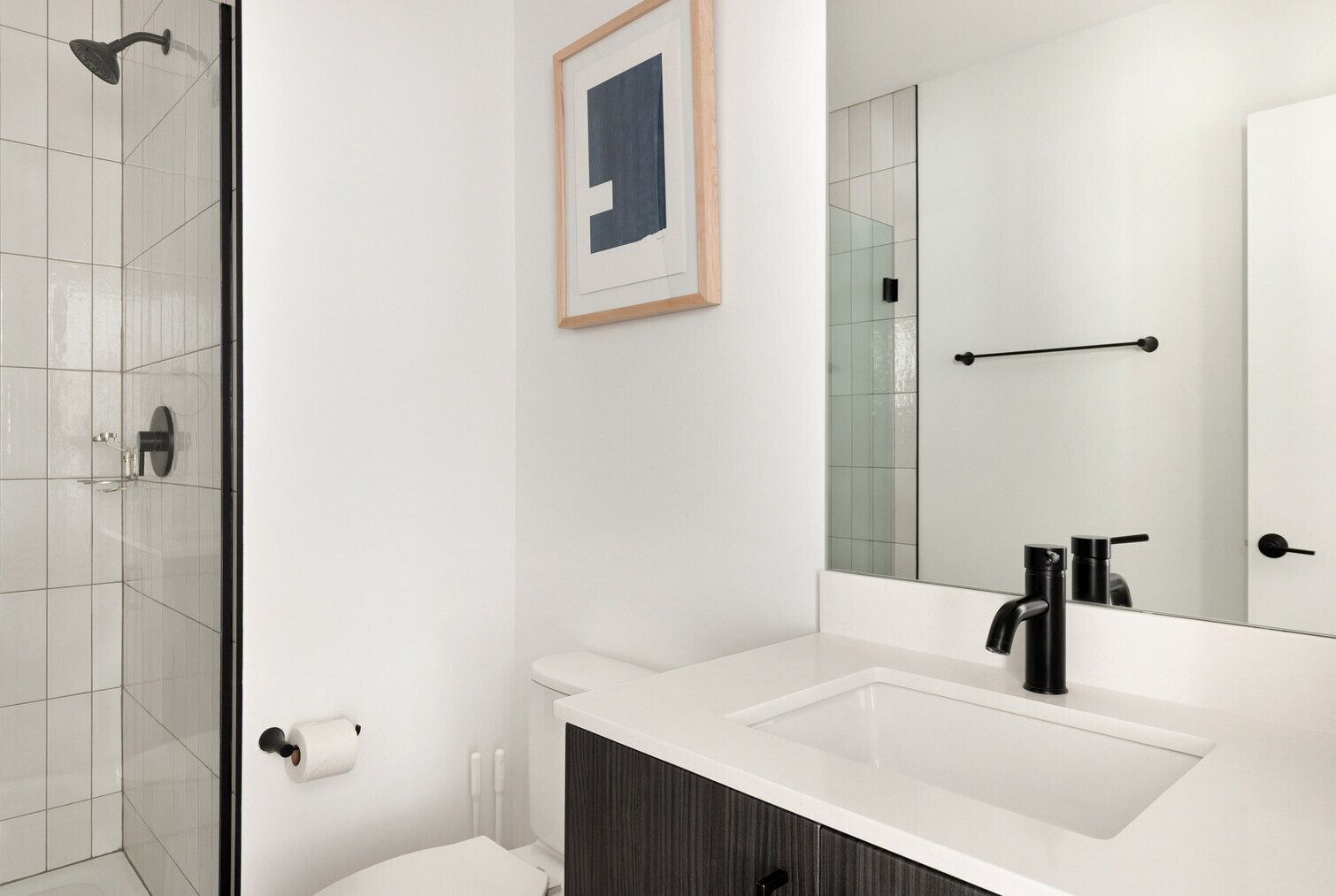 Un baño moderno con cabina de ducha de vidrio, un lavabo blanco con grifo negro y una obra de arte enmarcada en la pared.