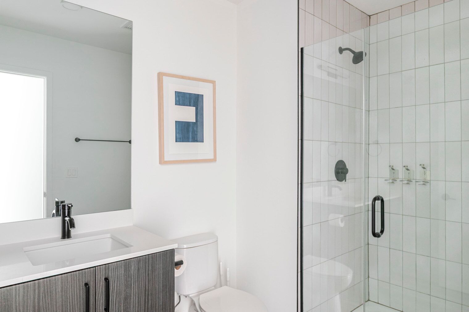 A modern bathroom with a large mirror, a framed artwork, a toilet, and a glass shower enclosure.