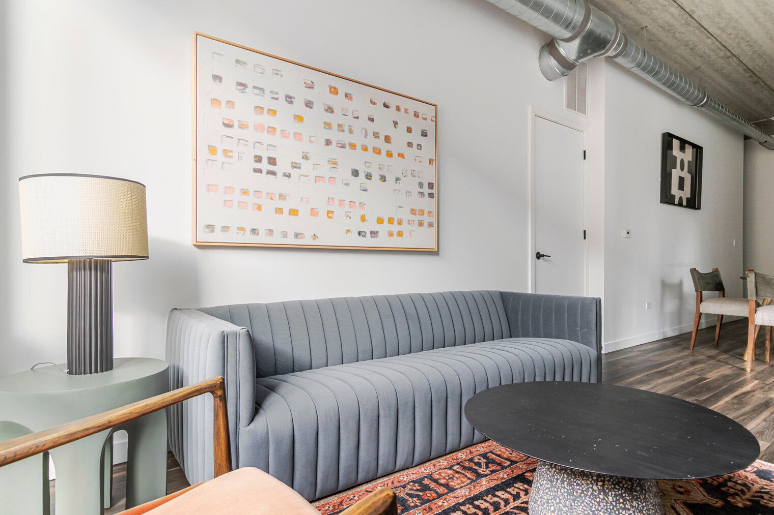 Una sala de estar moderna con un sofá gris, una mesa de centro redonda y una lámpara. Hay un cuadro abstracto grande en la pared y una puerta al fondo.