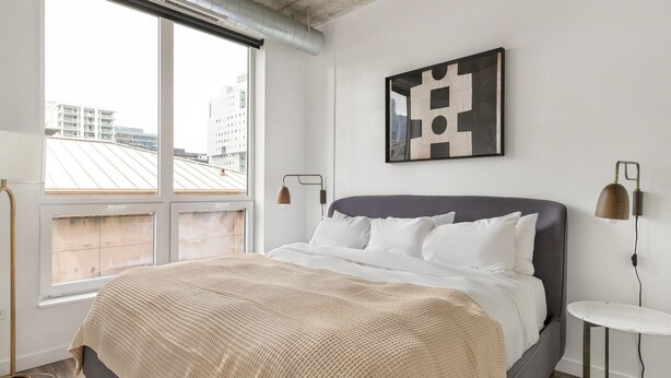 A modern bedroom with a large bed, a view of buildings through the window, and a geometric artwork on the wall. - Blueground | Near North Side, rooftop, nr Chicago River (Chicago)