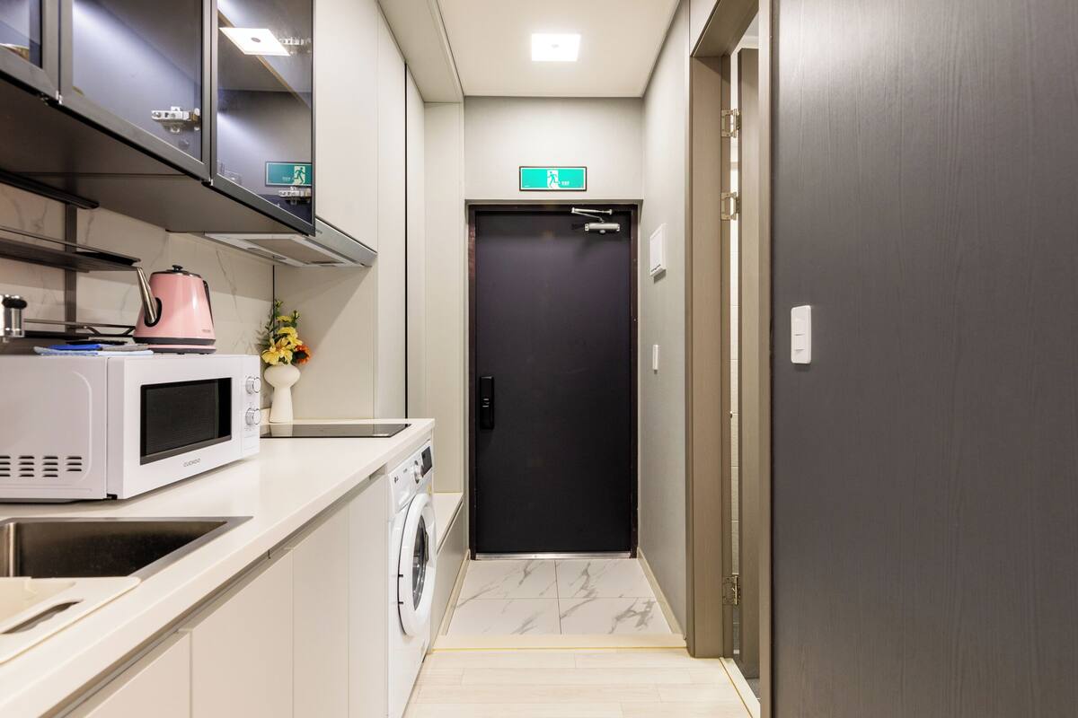 A narrow hallway with a microwave, washing machine, and cabinets on one side, and a dark door at the end.