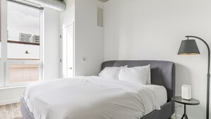 Ein modernes Schlafzimmer mit einem großen Bett, einem Nachttisch, einer Lampe und Blick auf Gebäude durch ein Fenster.