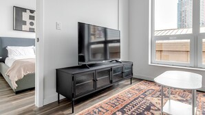 Ein modernes Schlafzimmer mit einem Bett, einem Fernseher auf einer schwarzen Kommode, einem kleinen weißen Tisch und einem großen Fenster mit Blick auf die Stadt.