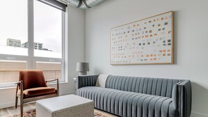 Ein modernes Wohnzimmer mit einem grauen Sofa, einem braunen Sessel, einem Couchtisch aus Beton und einem gemusterten Teppich. Ein großes abstraktes Gemälde hängt an der Wand.