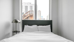 Ein Hotelzimmer mit Bett, Nachttisch, Lampe und Blick auf eine Stadtlandschaft durch große Fenster.