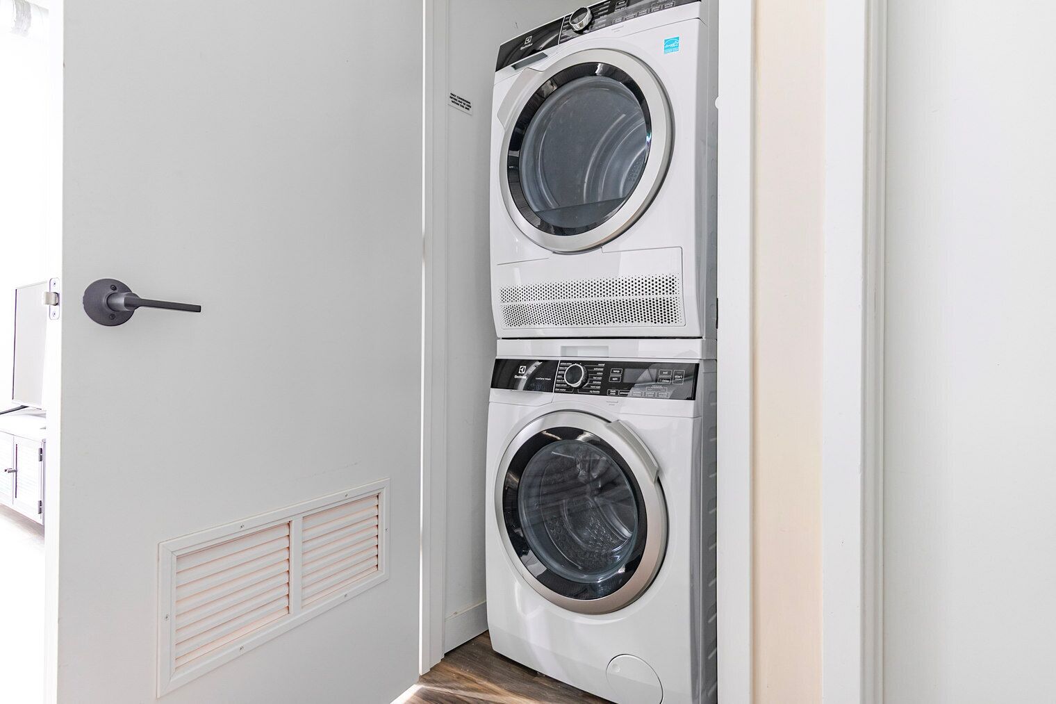 Ett kompakt tvättrum med två tvättmaskiner placerade ovanpå varandra, en dörr med handtag och en ventilationsöppning på väggen.
