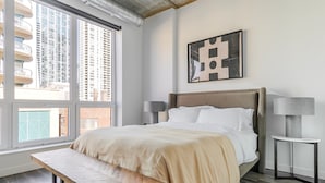 A modern bedroom with a large window, a bed with white linens, a bench, two bedside tables with lamps, and a geometric artwork on the wall. - Blueground | Near North Side, rooftop, nr Chicago River (Chicago)