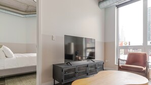 Een moderne hotelkamer met een bed, een televisie op een zwarte kast, een houten bijzettafeltje en een stadszicht door een groot raam.